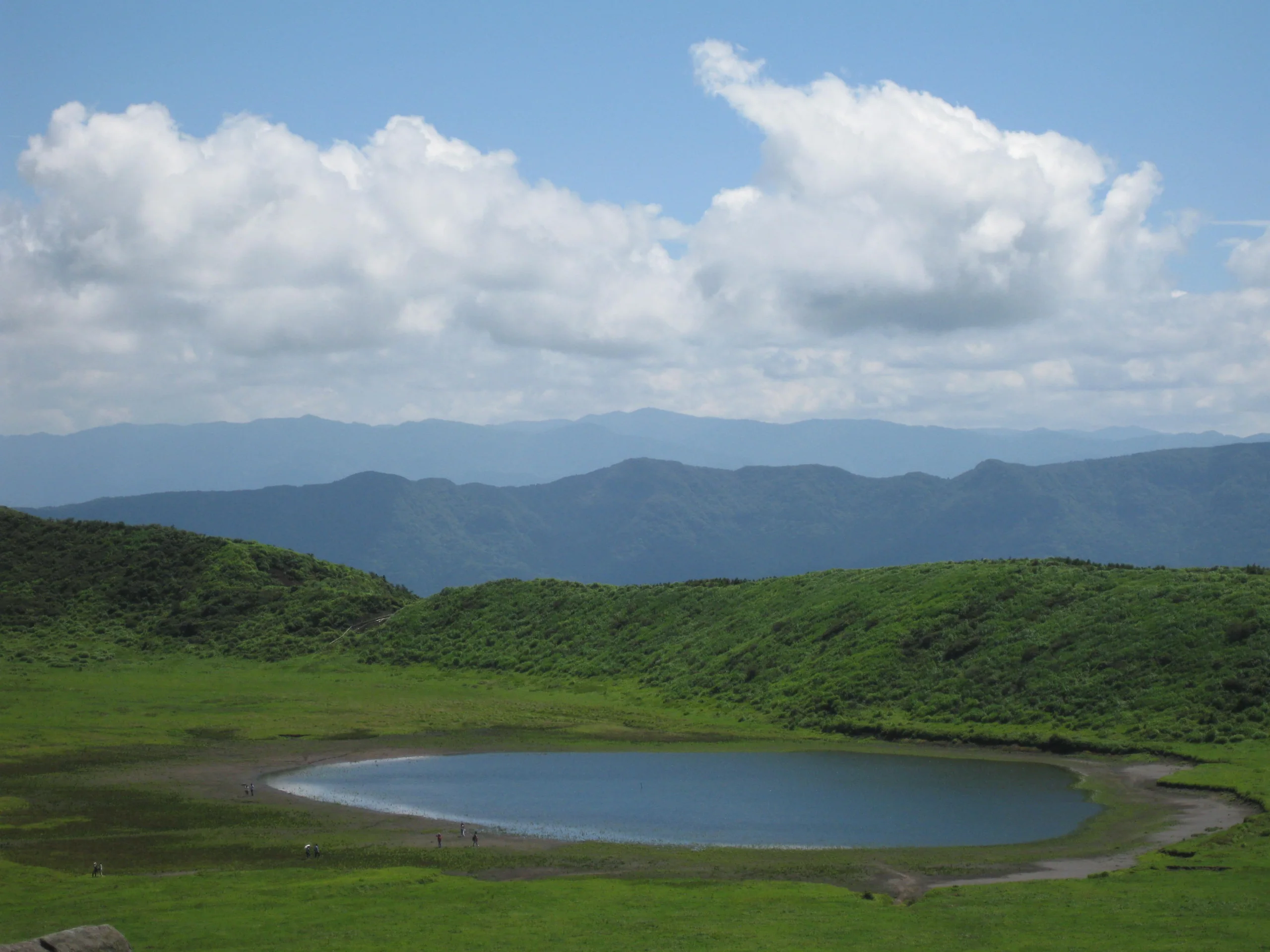 草千里草原環繞中央池塘，遠處是阿蘇山稜