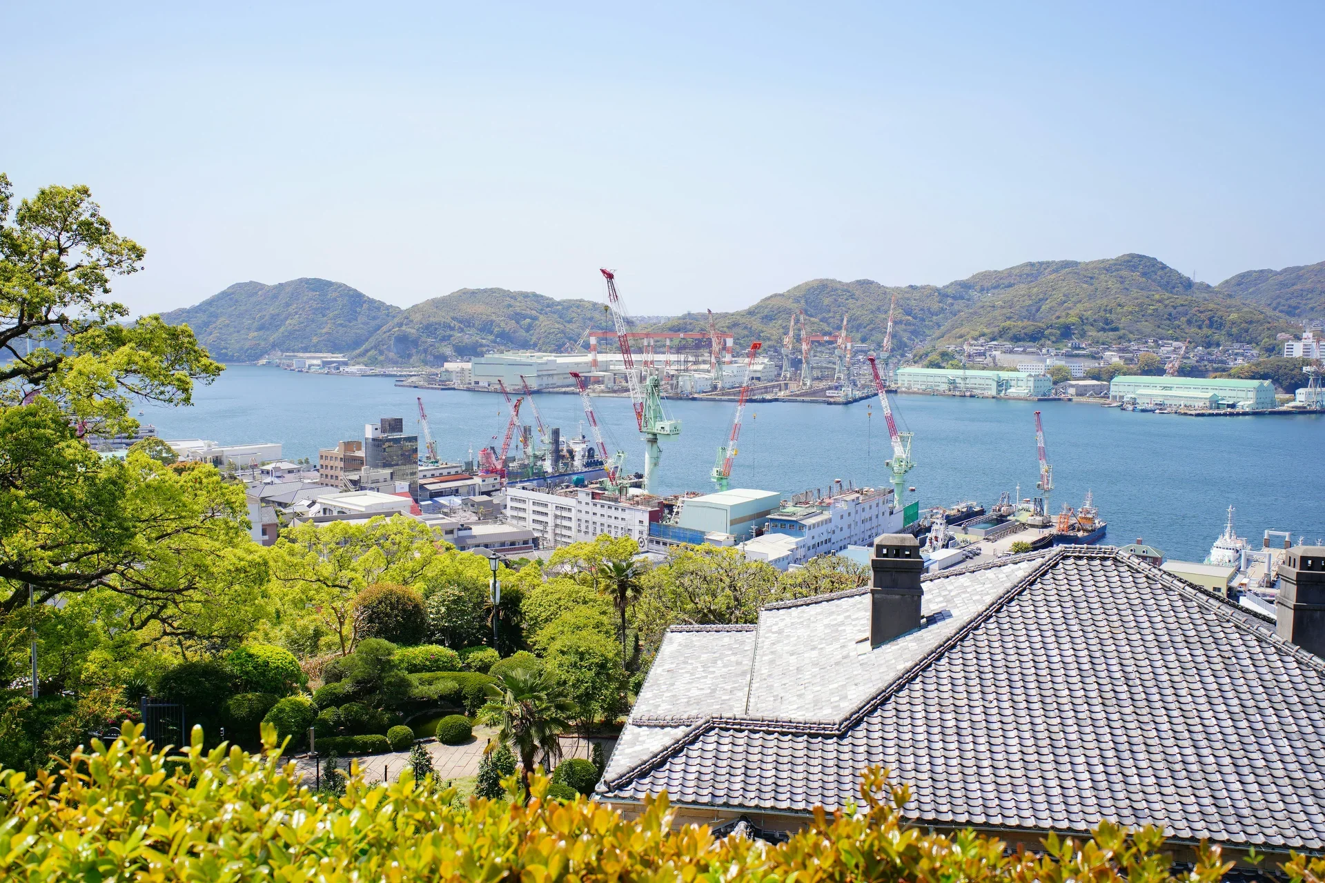 哥拉巴園俯瞰長崎港與造船廠起重機,前景是傳統瓦屋頂和園內綠樹